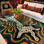 A modern living room scene featuring a large area rug with a bold abstract pattern in teal, orange, black, and cream. The rug is styled under a clear glass-top coffee table, next to a white sofa with patterned pillows and a leopard-print armchair.