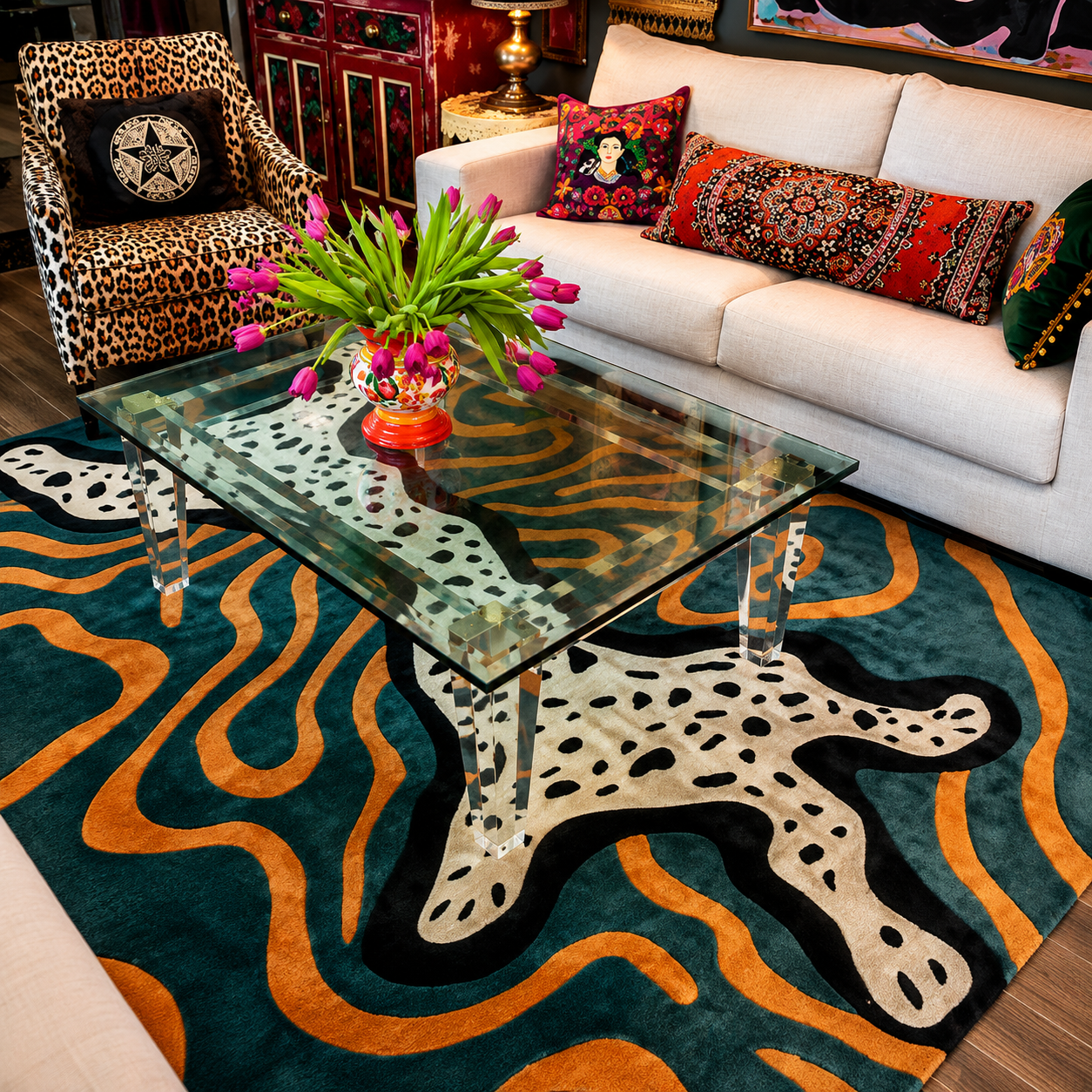 A modern living room scene featuring a large area rug with a bold abstract pattern in teal, orange, black, and cream. The rug is styled under a clear glass-top coffee table, next to a white sofa with patterned pillows and a leopard-print armchair.