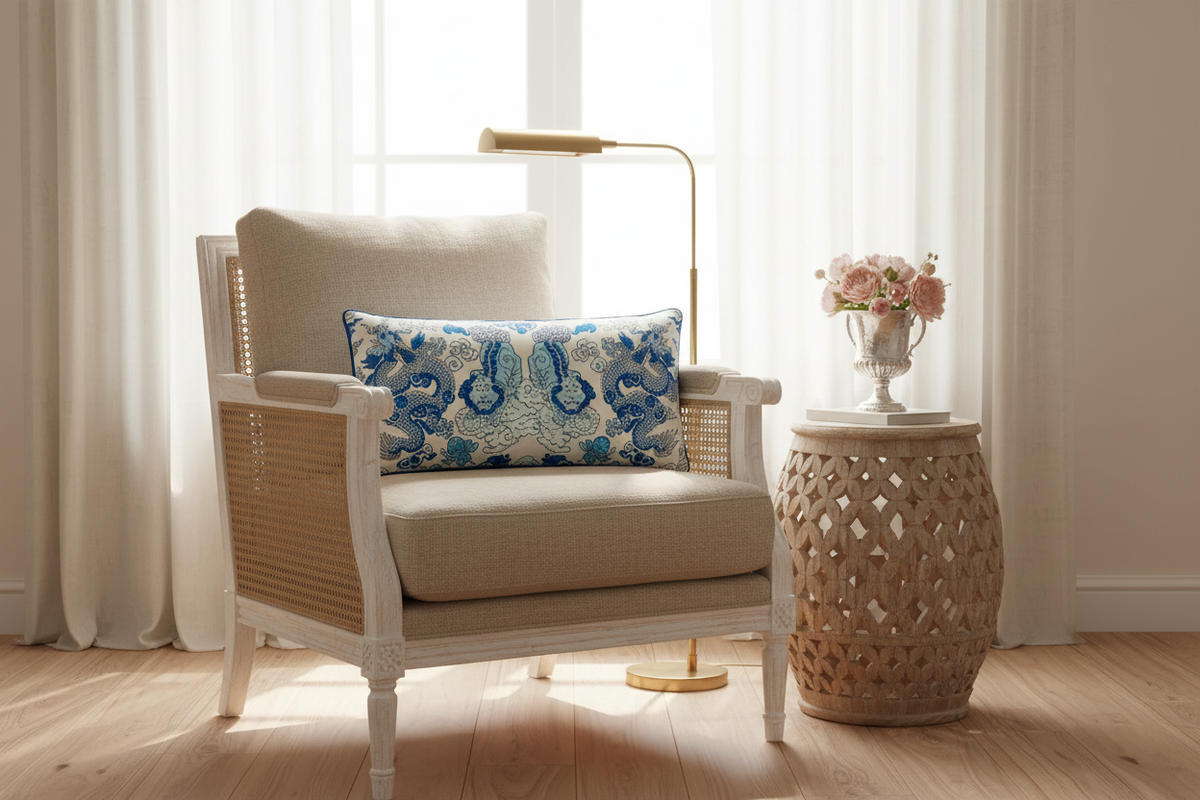 A styled interior scene with a beige upholstered armchair featuring woven cane sides, a blue and white patterned accent pillow, a gold floor lamp, a carved wood side table, and a vase of pink flowers in front of sheer curtains.