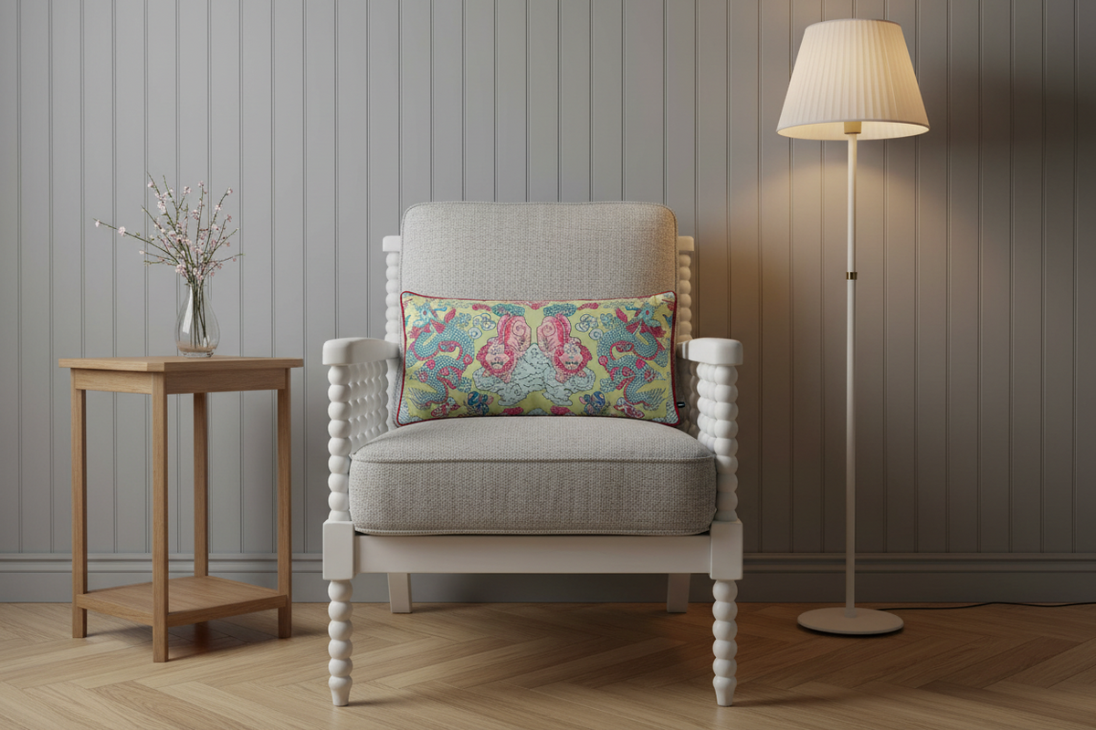 A light gray upholstered armchair with turned white wooden arms and legs, styled with a colorful paisley accent pillow. The chair is set in a cozy room with a light wood side table on the left and a white floor lamp on the right, against a gray paneled wall.
