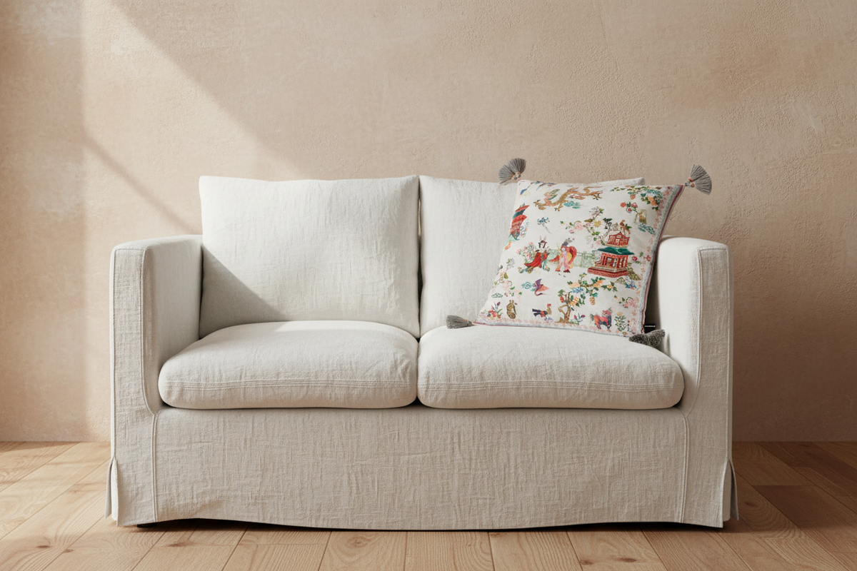A cream-colored linen slipcovered loveseat sofa with two seat cushions and two back cushions, styled with a decorative patterned accent pillow, set against a textured beige plaster wall and light wood flooring.