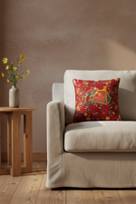 A white wooden armchair with woven cane detailing and beige cushions, styled in a bright room with sheer white curtains, a patterned red throw pillow, a gold floor lamp, and a decorative side table.