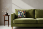 An olive green velvet sofa in a styled room setting, featuring a decorative throw pillow with a colorful Japanese ukiyo-e woodblock print and a small wooden side table with dried branches on a textured light gray wall.