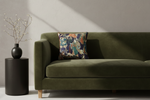 An olive green velvet sofa in a modern room setting, styled with a square throw pillow featuring a detailed Japanese ukiyo-e woodblock print. A black cylindrical side table with a black vase of branches stands to the left against a textured off-white wall with dramatic diagonal sunlight casting shadows.