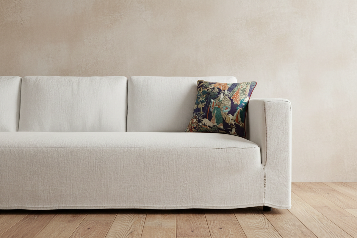A white fabric slipcover on a three-seater sofa, styled with a decorative multicolor patterned accent pillow, set against a textured beige wall and light wood flooring.