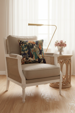 A classic outdoor-style armchair with a white frame and woven cane backrest, styled with a beige cushion and a decorative multicolor patterned throw pillow. It is set in a bright room next to a wooden side table with a vase of pink flowers and a gold floor lamp.