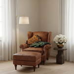 An olive green velvet sofa in a styled room setting, featuring a tiger-patterned throw pillow and a green crocodile plush toy. A black cylindrical side table with a matching vase holding dried branches stands to the left against a beige wall with soft diagonal shadows.