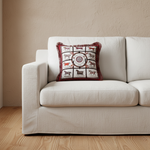 A cream slipcovered sofa in a warm, rustic room setting, featuring a decorative throw pillow with a red and white horse-themed pattern. A small wooden side table with a vase of yellow flowers sits to the left.