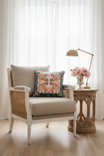 A cream-colored armchair with woven cane details and a decorative multicolor patterned throw pillow, styled in a bright room next to a carved wooden side table with a lamp and pink flowers.
