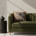 An olive green velvet loveseat sofa in a styled room setting, featuring a decorative throw pillow with a multicolor leopard print pattern and gold fringe. A black side table with a black vase is positioned to the left.