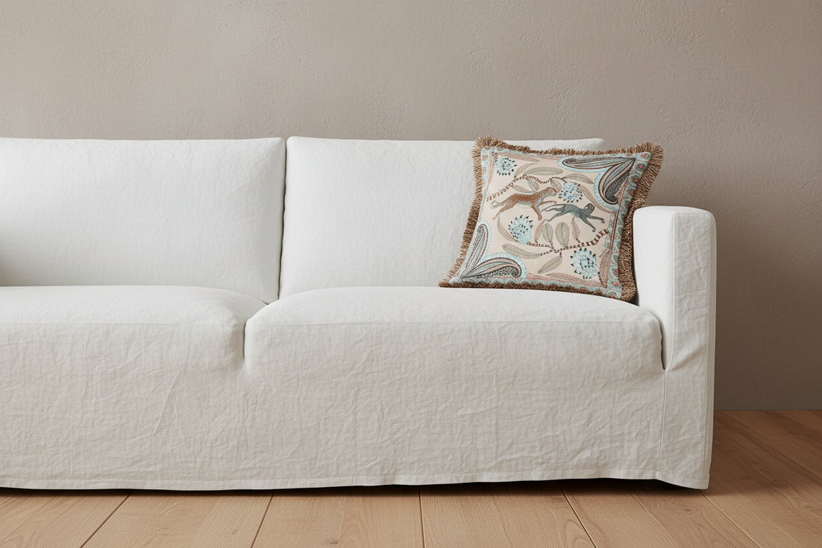 A white slipcovered sofa with a decorative beige throw pillow featuring blue and brown floral patterns, set against a neutral beige wall and light wood floor.