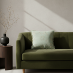A cream-colored slipcovered armchair with a light green textured accent pillow, set against a warm brown plaster wall with a wooden side table on the left.