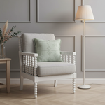 A cream-colored slipcovered armchair with a light green textured accent pillow, set against a warm brown plaster wall with a wooden side table on the left.
