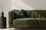 An olive green velvet sofa with vertical channel tufting in a styled room, featuring a dark teal floral throw pillow and a black side table with a vase.