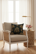 A styled interior scene with a white and wicker armchair in a bright room, featuring a dark green throw pillow with a gold palm leaf pattern. A carved wooden side table next to the chair holds a gold floor lamp and a vase of pink flowers.