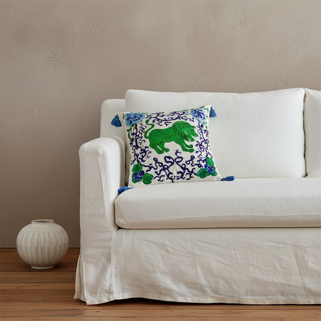 A decorative throw pillow with a green elephant and blue floral pattern on a white background, styled on an off-white slipcovered sofa next to a small white ceramic vase against a textured beige wall.