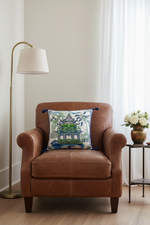 A brown leather armchair in a bright room, styled with a square decorative throw pillow featuring a green pagoda and tropical foliage pattern. A brass floor lamp stands to the left, and a small side table with white flowers is to the right.