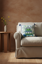 A brown leather armchair in a bright room, styled with a square decorative throw pillow featuring a green pagoda and tropical foliage pattern. A brass floor lamp stands to the left, and a small side table with white flowers is to the right.