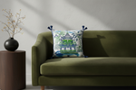 A brown leather armchair in a bright room, styled with a square decorative throw pillow featuring a green pagoda and tropical foliage print. A brass floor lamp stands to the left, and a small side table with white flowers is to the right.