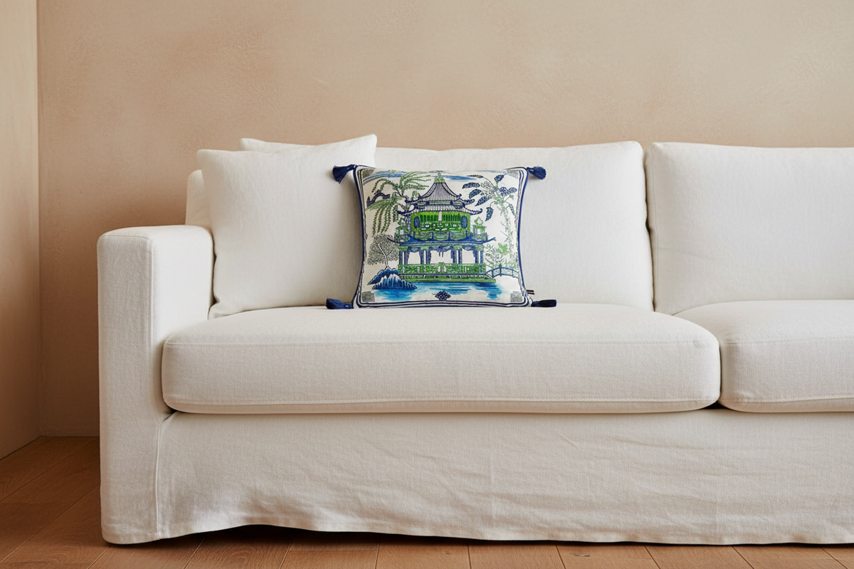 A brown leather armchair in a bright room, styled with a square decorative throw pillow featuring a green pagoda and tropical foliage pattern. A brass floor lamp stands to the left, and a small side table with white flowers is to the right.