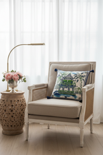A brown leather armchair in a bright room, styled with a square decorative throw pillow featuring a green pagoda and tropical foliage pattern. A brass floor lamp stands to the left, and a small side table with white flowers is to the right.