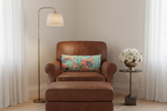 A brown leather armchair with a matching ottoman in a styled room setting, featuring a colorful floral throw pillow, a brass floor lamp, and a side table with white flowers.
