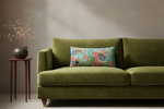 An olive green velvet sofa in a styled room setting, featuring a colorful rectangular throw pillow with a tropical floral print, next to a small wooden side table with dried branches.