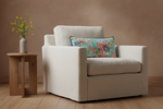 A cream-colored upholstered armchair with a colorful floral throw pillow, set in a room with a textured brown wall and light wood flooring, next to a small wooden side table with a vase of yellow flowers.