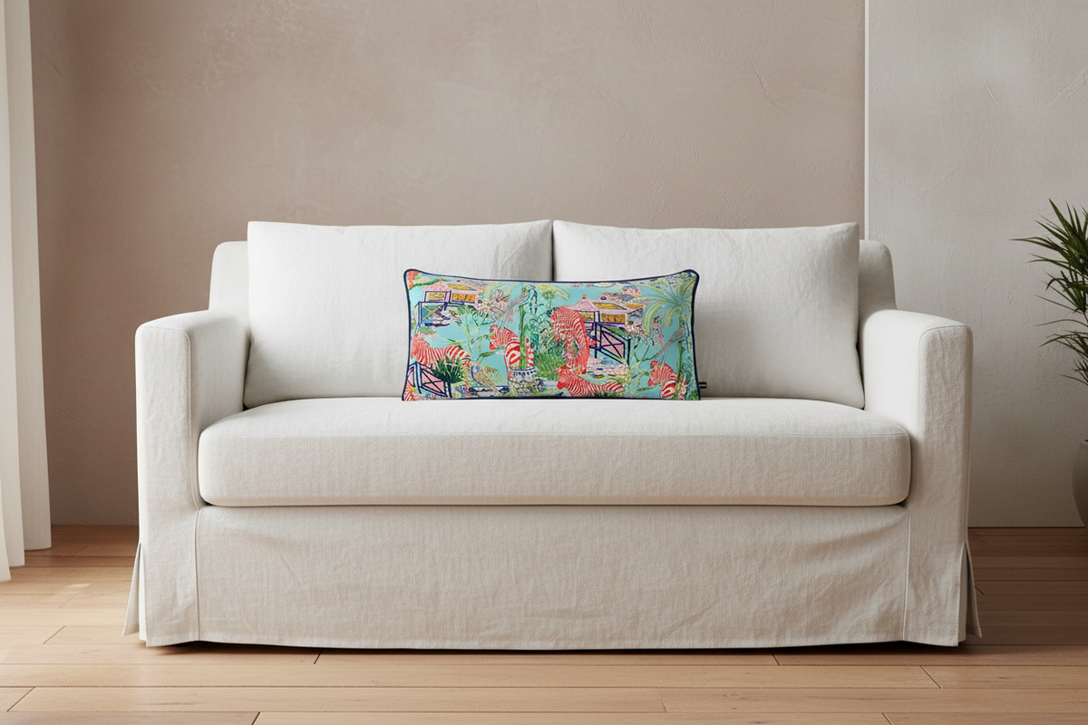 A cream-colored slipcovered loveseat sofa with a colorful abstract patterned lumbar pillow, set against a neutral beige wall and light wood floor.