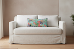 A cream-colored slipcovered loveseat sofa with a colorful abstract patterned lumbar pillow, set against a neutral beige wall and light wood floor.