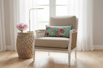A beige upholstered armchair with a colorful tropical floral throw pillow, styled in a bright room next to a carved wooden side table with pink flowers and a gold floor lamp.