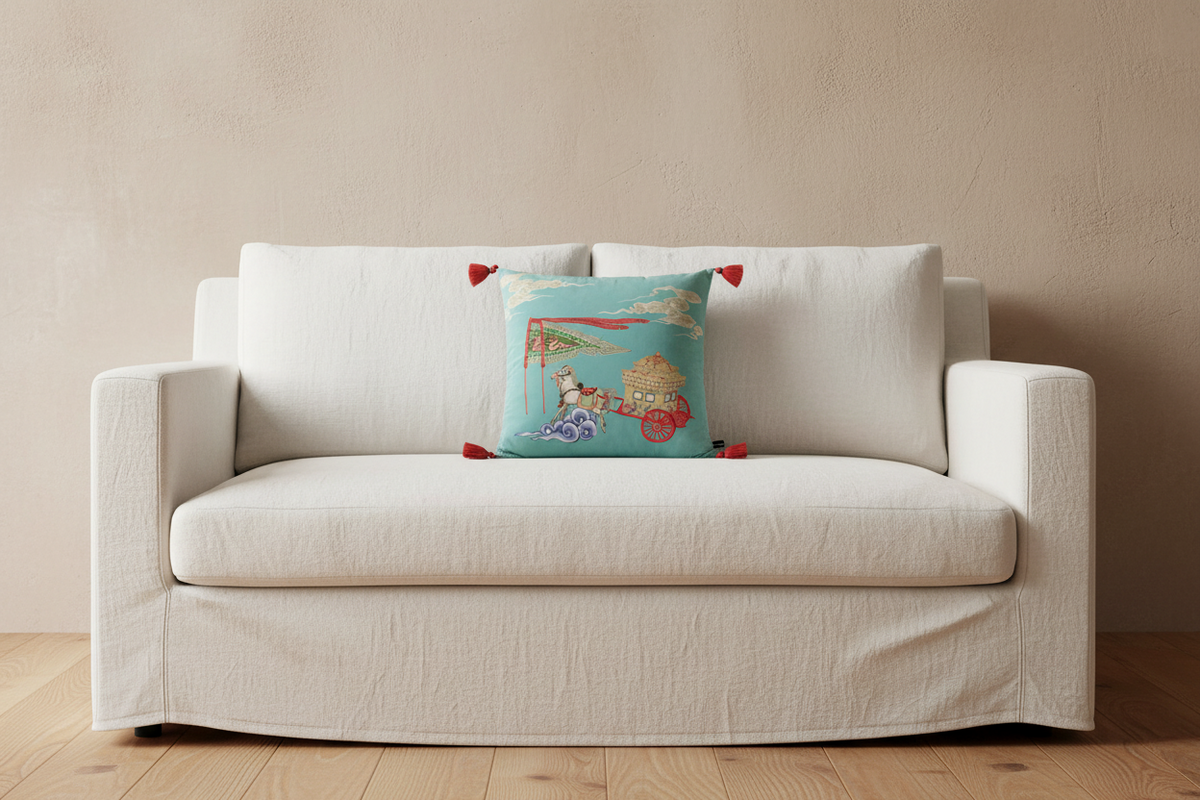 A cream-colored slipcovered loveseat sofa with a decorative square throw pillow featuring a colorful Japanese-style illustration and red tassels, set against a textured beige wall and light wood floor.