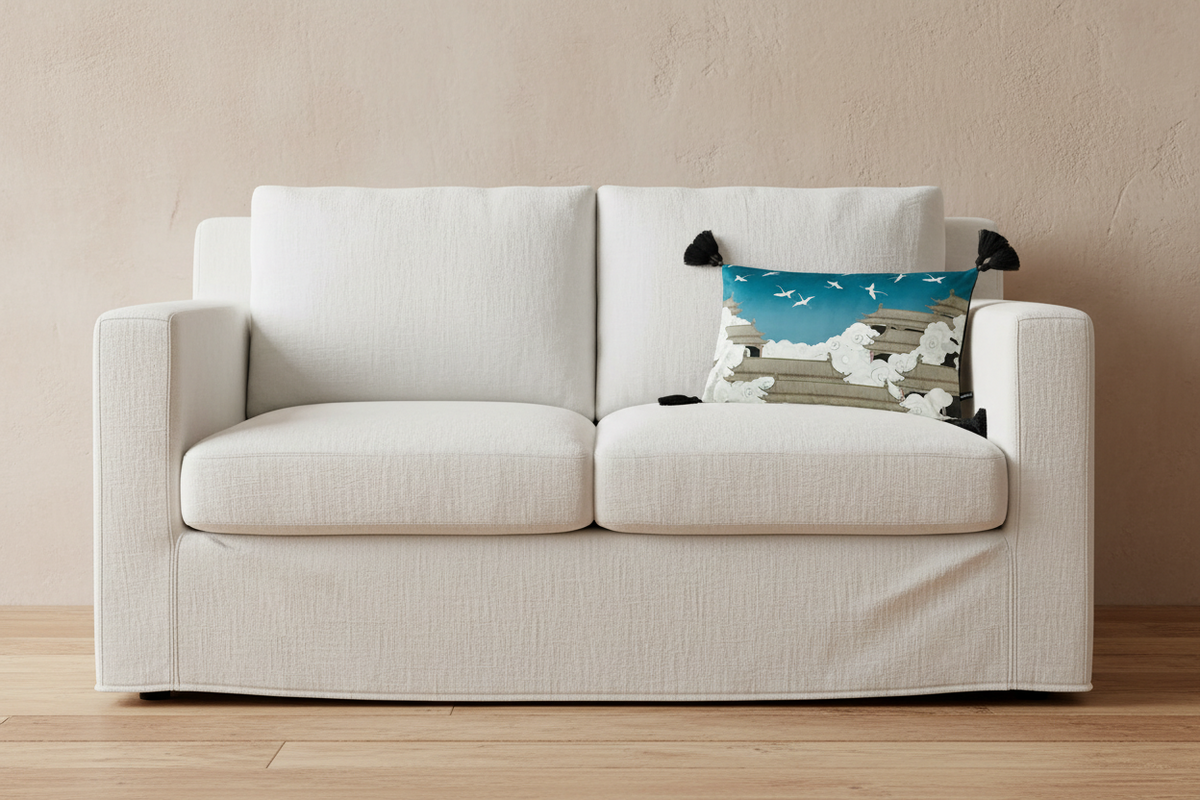 A modern two-seater sofa upholstered in textured off-white fabric, set against a warm beige wall and light wood floor. A decorative throw pillow with a blue sky and white clouds design rests on the right side of the sofa.