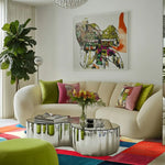 A vibrant and artistic area rug enhances a modern living room setting. The hand-tufted rug displays an abstract cityscape design, bringing a splash of color with its rich hues. Perfect for large living rooms, dining rooms, or as stylish dining room carpets, this wool rug complements the contemporary decor, including a plush sofa and metallic coffee table.