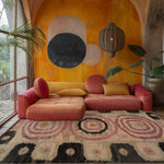 A vibrant living room featuring the Blossom Mandala Hand Tufted Rug. This area rug, perfect for dining room areas, showcases an intricate mandala design in pink and black tones. The wool rug adds warmth and style, complementing the colorful couch and bold wall art.