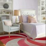 A stylish bedroom setting featuring the Blushing Pinwheel Hand Tufted Rug with a vibrant pink and red swirling pattern. The area rug adds a pop of color to the cozy space, making it perfect for both living room and office settings. The elegant wool rug complements the plush armchair and sophisticated décor, creating a warm and inviting atmosphere.