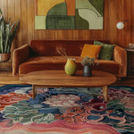 A vibrant Coral Bloom Hand Tufted Rug adds a splash of color to a cozy living room. The area rug features floral and abstract patterns in shades of blue, pink, and orange, perfectly complementing the warm tones of the wooden decor and plush sofa. Ideal for those seeking wool rugs or area rugs for living rooms, especially in an 8x10 size.