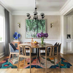 A vibrant and abstract Cosmic Tides Hand Tufted Area Rug elevates the living room space with its colorful design. Perfect for enhancing large area rugs in a sophisticated setting, the wool rug sits beautifully beneath a round dining table surrounded by modern chairs. The room is adorned with elegant decor, including a striking chandelier and lush purple flowers, making it an ideal choice for those seeking stylish rugs for living room aesthetics.