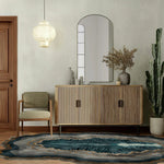 Stylish living room featuring the Crystal Depths Hand Tufted Viscose Area Rug with abstract, swirling patterns in blue and grey, placed on wooden flooring. The room is elegantly decorated with a modern wooden sideboard, a comfortable chair, and a hanging light fixture, creating a cozy and inviting atmosphere.