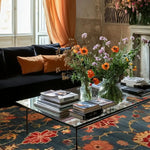 A cozy living room featuring a large, hand-knotted area rug with a vibrant floral design. The square rug, perfect for a vintage touch, is placed under a glass coffee table adorned with books and flowers, enhancing the warm ambiance of the space. Ideal for both living and dining rooms, this wool rug adds a Persian-inspired elegance.