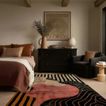 Stylish living room featuring a Dusky Petals Hand Tufted Area Rug. The large wool rug, with its abstract design in pink and black tones, complements the cozy brown bedding and modern black furniture, creating a warm and inviting atmosphere ideal for any living room.