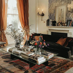 A cozy living room featuring the Egyptian Enigma Wool Hand Knotted Area Rug. This vintage rug showcases intricate patterns and rich colors, adding warmth and character to the space. Perfect for those seeking elegant rugs for living room decor, this area rug complements a stylish black sofa and a glass coffee table adorned with books and flowers.