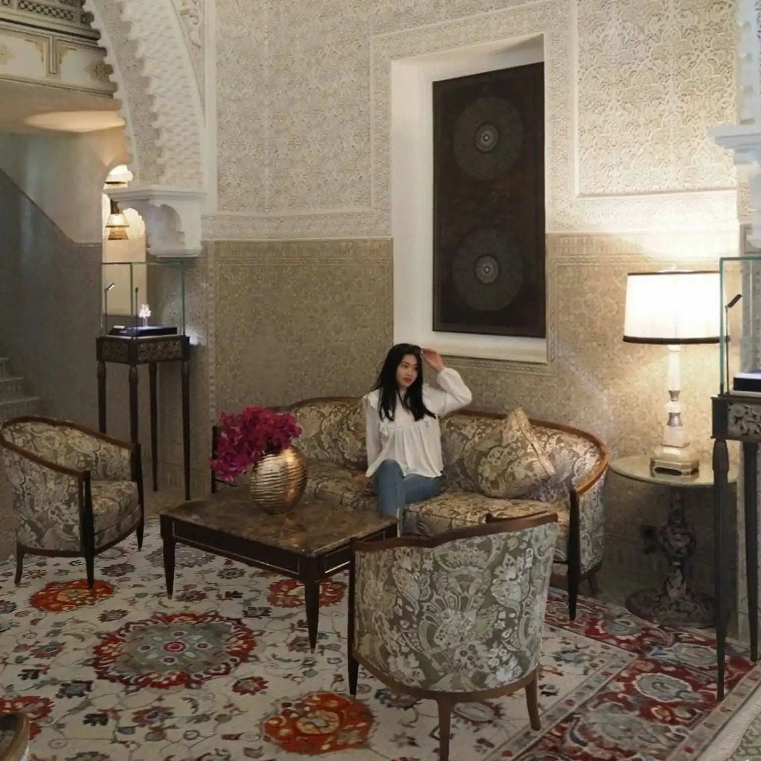 Woman sitting on a couch in an ornate room with intricate floral persian rug in red and beige color, wall patterns and decorative furniture.