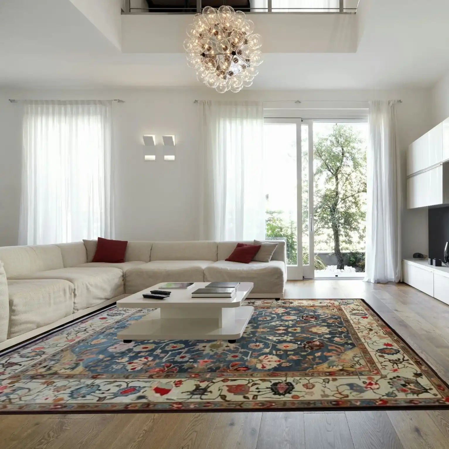 Elegant living room featuring a large, blue floral hand-knotted area rug with intricate patterns, complementing a modern white sectional sofa and a chic coffee table, under a stylish chandelier.