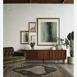 A beautifully styled living room featuring a large Elephant Whisper hand-tufted wool rug. The area rug, perfect for living rooms, showcases an intricate design with a central elephant motif. It sits on a wooden floor, complementing the mid-century modern furniture, including a plush green sofa and a sleek wooden sideboard adorned with framed art and greenery.