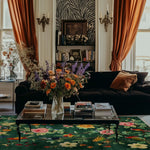Elegant living room featuring the Emerald Oasis Hand Tufted Area Rug. The vibrant wool rug with floral patterns enhances the space, complemented by a plush black sofa and a coffee table adorned with books and a colorful flower arrangement. Perfect for living room and dining room settings.