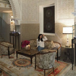 Woman sitting on a couch in an ornate room with intricate beige handmade rug, wall patterns and decorative furniture.