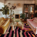 Stylish living room featuring an Evolving Shapes Hand Tufted Area Rug with a vibrant abstract pattern in pink, black, and yellow. The wool rug complements the plush velvet sofas, creating a cozy and inviting ambiance. Ideal for living room and dining room settings, this formal dining room rug adds a touch of elegance to the space, enhanced by lush green plants and a modern glass coffee table.