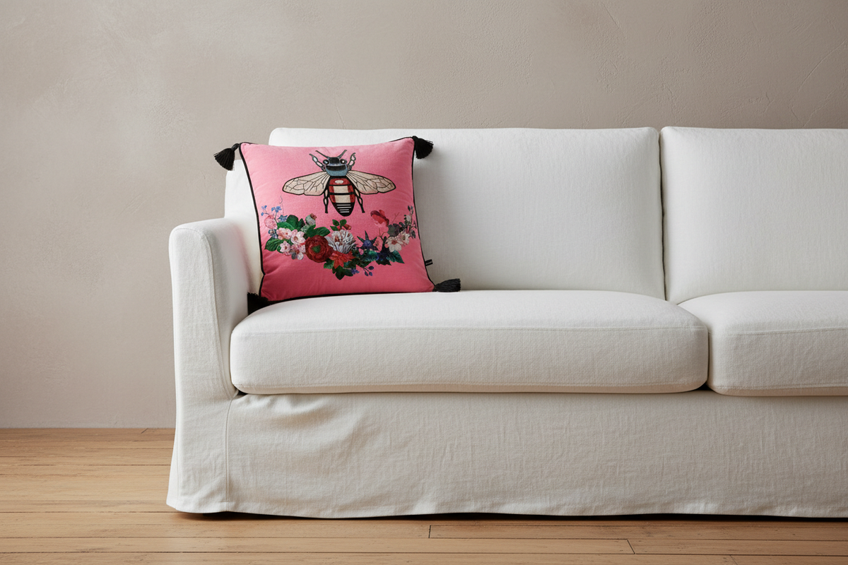 A pink decorative throw pillow featuring a detailed bee and floral embroidery, styled on a white slipcovered sofa against a neutral beige wall and light wood floor.