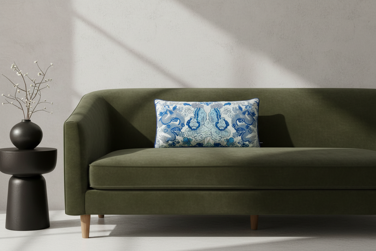 A white armchair with woven cane panels and a beige upholstered seat, styled with a blue and white patterned accent pillow. It is set in a bright room with white sheer curtains, a wooden floor, a matching woven side table, a gold floor lamp, and a vase of pink flowers.